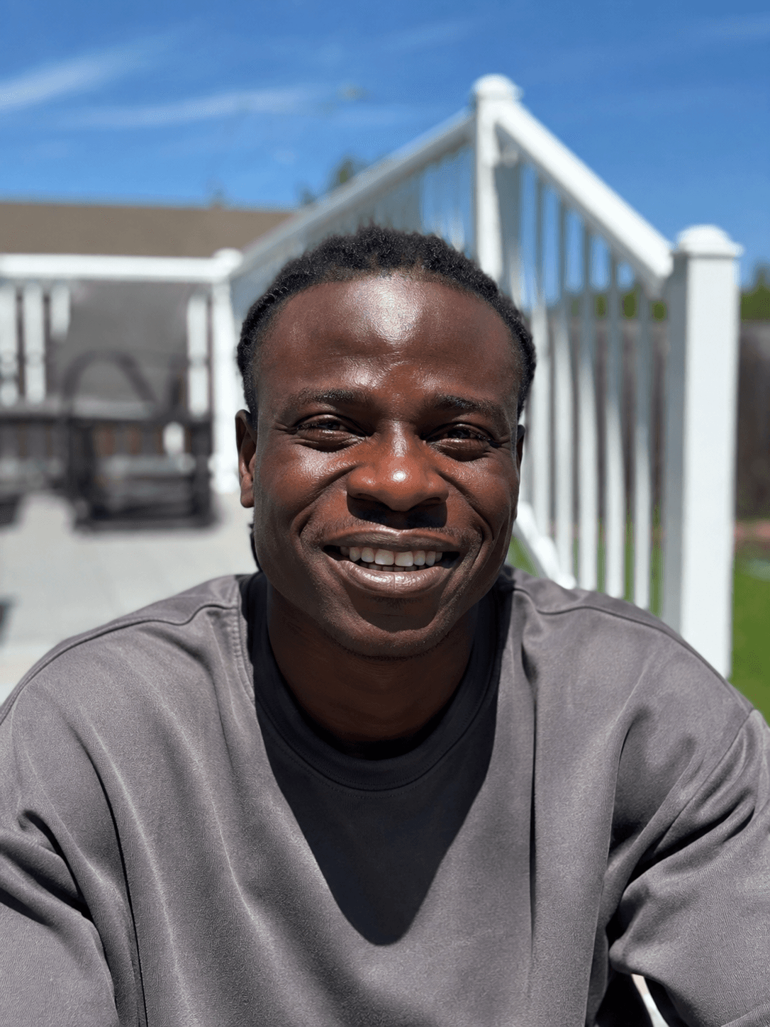 Smiling Black man in a grey sweatshirt sits outdoors on a sunny deck with railings.